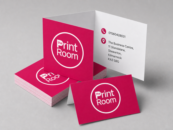 Folded business cards featuring The Print Room round magenta logo on the front, with the phone number and business address printed inside, displayed alongside a stack of matching premium business cards on a neutral grey studio background.