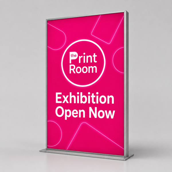Freestanding LED lightbox display with a silver frame featuring The Print Room round magenta logo, the message "Exhibition Open Now," and an illuminated pink graphic on a neutral grey studio background.