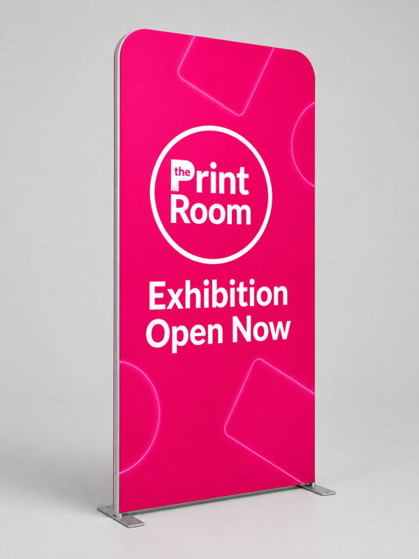 Stretch fabric exhibition display featuring The Print Room round magenta logo, the message "Exhibition Open Now," a vibrant pink graphic with subtle geometric accents, and a freestanding fabric display on a neutral grey studio background.