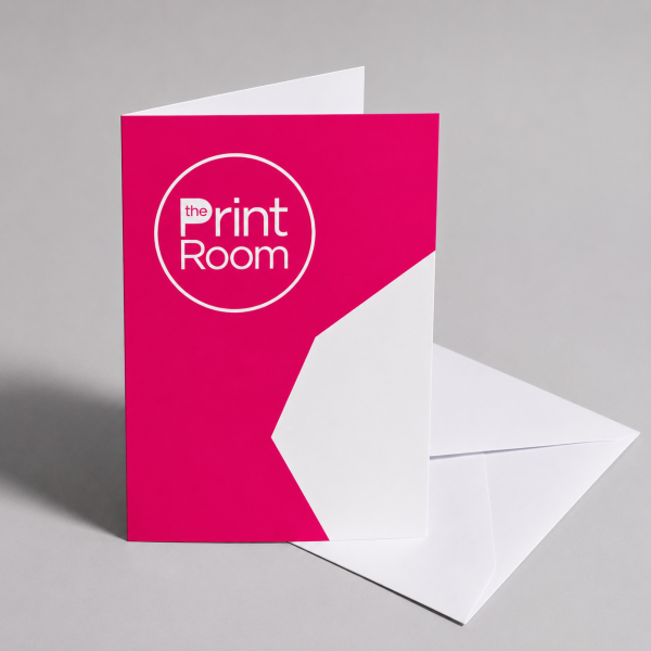 Folded greeting card with matching white envelope, featuring the Print Room circular logo and vibrant pink branded design, displayed on a light grey studio background.
